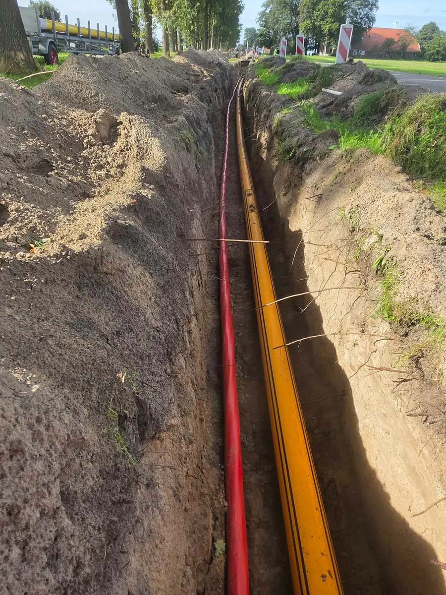 Open sleuf langs de weg met een rode waterleiding en een gele gasleiding erin